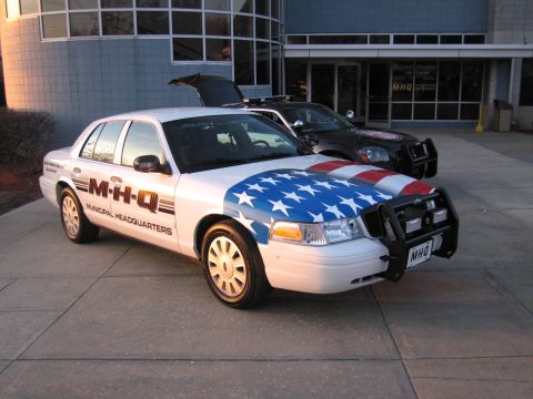 1991 Ford LTD Crown Victoria - white Ford Crown Victoria with an American-flag hood wrap, push bar, and M-H-Q Municipal Headquarters livery parked outside a building.