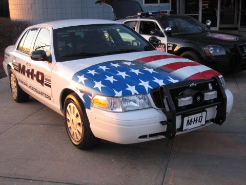 1991 Ford LTD Crown Victoria - Front three-quarter view of a white Crown Victoria with US-flag hood graphic, black push bar, and M-H-Q lettering, hood raised.