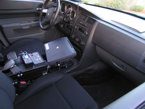1991 Ford LTD Crown Victoria - Interior of a police vehicle showing a center-console laptop mount, two-way radio, gear shifter, and the dashboard gauge cluster.