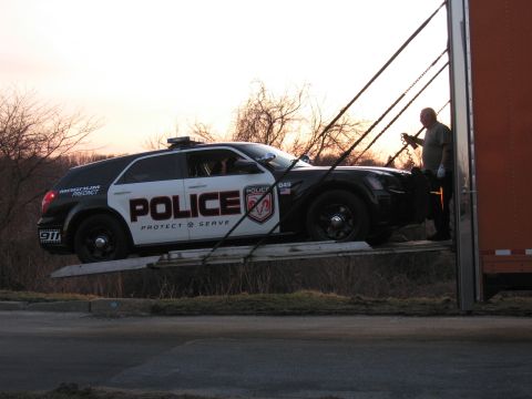 1991 Ford LTD Crown Victoria - man loads a black-and-white POLICE Dodge Magnum wagon up a ramp into an enclosed trailer at sunset.