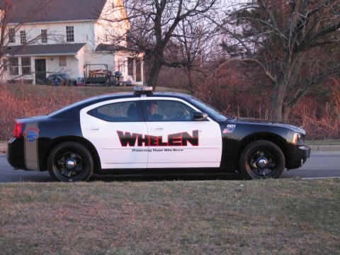 1991 Ford LTD Crown Victoria - black-and-white Dodge Charger sedan with large red WHELEN 'Protecting Those Who Serve' decals parked on a roadside at dusk.