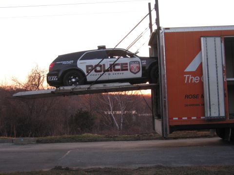 1991 Ford LTD Crown Victoria - black-and-white POLICE-marked Dodge SUV sits on the raised ramp of an orange enclosed auto transport trailer at sunset.