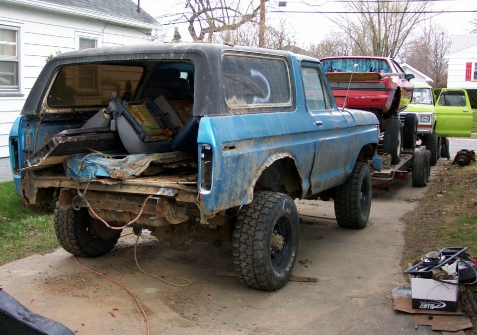 1978 Ford Bronco on a trailer, being towed. Another vehicle is on a trailer behind it, and a green truck is parked next to it