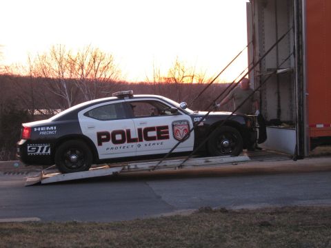 1991 Ford LTD Crown Victoria - black-and-white POLICE Dodge Charger rests on the ramp of an enclosed auto carrier as a worker stands at the trailer opening at sunset.