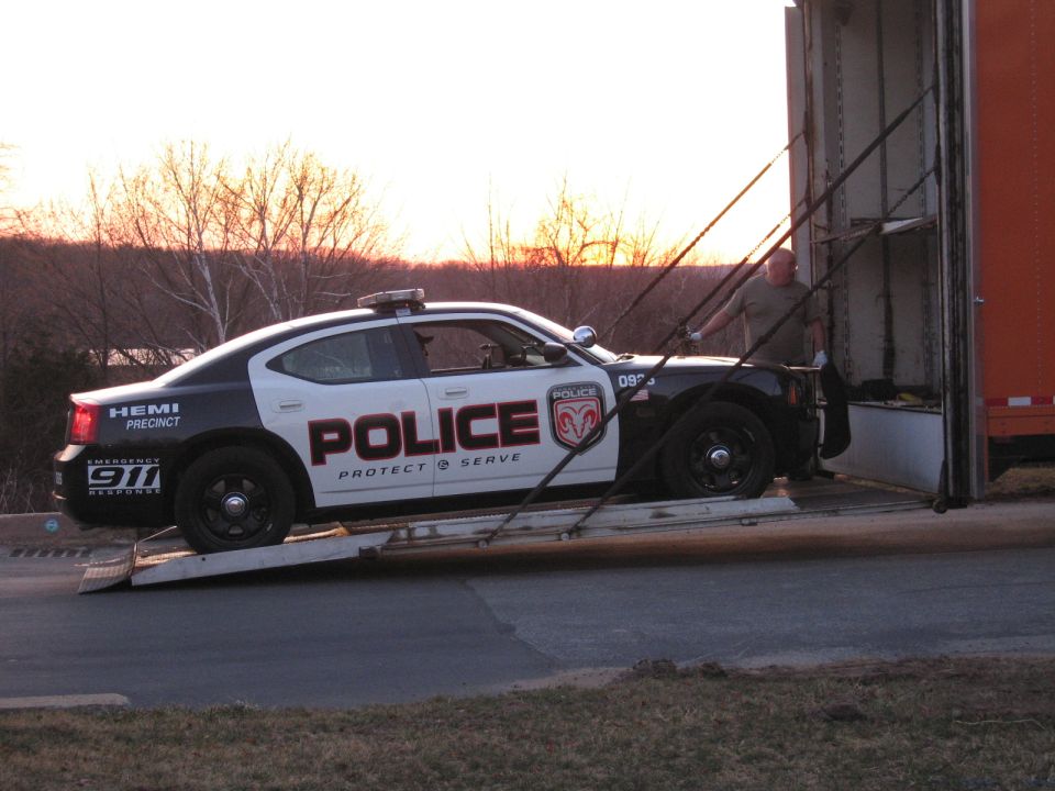 Police Dodge Charger on an enclosed auto carrier ramp with a worker at sunset
