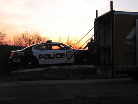 1991 Ford LTD Crown Victoria - black-and-white POLICE Dodge Charger on an auto-carrier ramp silhouetted against a bright orange sunset, a worker beside the trailer.
