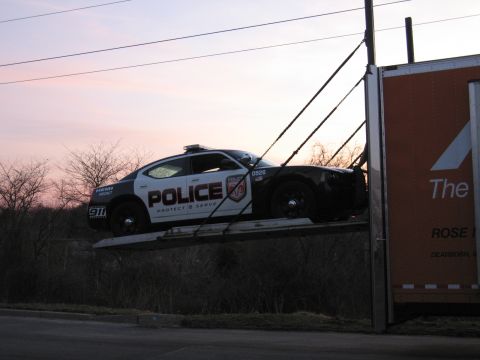 1991 Ford LTD Crown Victoria - black-and-white POLICE Dodge Charger raised high on the ramp of an orange enclosed auto-transport trailer at dusk.
