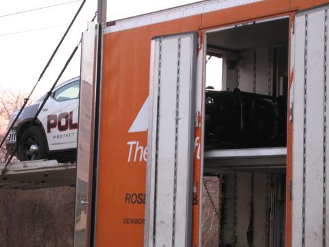 1991 Ford LTD Crown Victoria - open rear doors of an orange enclosed auto-transport trailer reveal a white POLICE car on the upper deck and a dark car below.