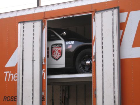 1991 Ford LTD Crown Victoria - Through the rear doors of an orange auto carrier, a white POLICE car numbered 0926 with a red Dodge Ram-head emblem sits loaded inside.