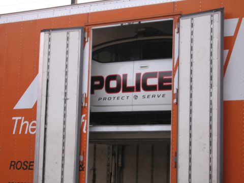 1991 Ford LTD Crown Victoria - Through the rear doors of an orange auto carrier, the rear of a white car marked POLICE 'Protect & Serve' is visible on the upper deck.