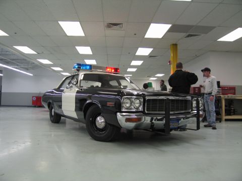 1991 Ford LTD Crown Victoria - 1970s-era black-and-white Dodge police sedan with roof light bar and front push bar displayed indoors as two men talk nearby.