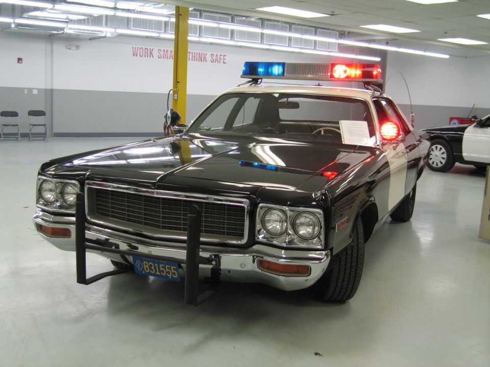 Black-and-white classic Dodge police car with light bar, front three-quarter view indoors