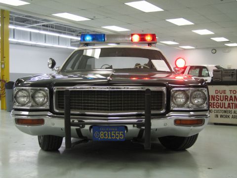 1991 Ford LTD Crown Victoria - Head-on view of a black-and-white Dodge police cruiser showing its chrome grille, dual round headlights and lit roof light bar.