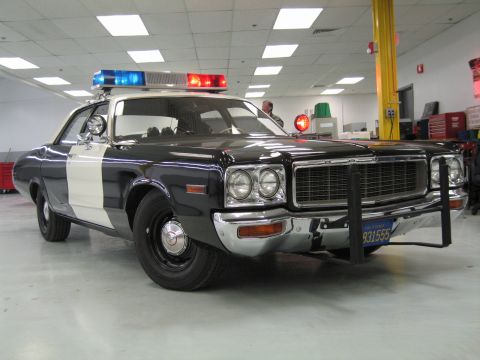 1991 Ford LTD Crown Victoria - vintage black-and-white police cruiser with white door panel and chrome push bumper, parked indoors with its light bar on.