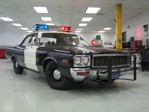 1991 Ford LTD Crown Victoria - black-and-white Dodge police cruiser, DODGE lettering on the grille, with spotlight and lit roof light bar parked indoors.