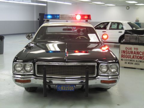 1991 Ford LTD Crown Victoria - Head-on view of a black-and-white police cruiser with chrome grille, push bumper and lit emergency lights near a warning sign.