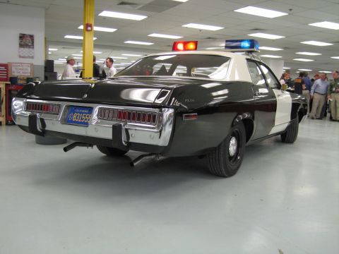 1991 Ford LTD Crown Victoria - Rear three-quarter view of a black-and-white police cruiser with lit roof light bar and full-width taillights among people in a hall.