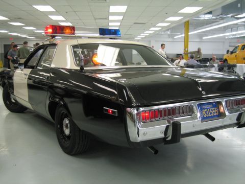 1991 Ford LTD Crown Victoria - Rear three-quarter view of a black-and-white police cruiser with lit light bar, a yellow truck and crowd visible in the background.