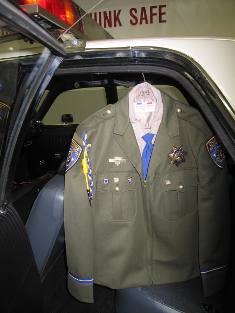 1991 Ford LTD Crown Victoria - CHP officer's olive uniform jacket with patches, badge, and blue tie hangs inside the rear of a black-and-white patrol car.