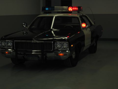 1991 Ford LTD Crown Victoria - black-and-white Dodge police car with quad round headlights and flashing roof and grille lights sits in a dim garage.