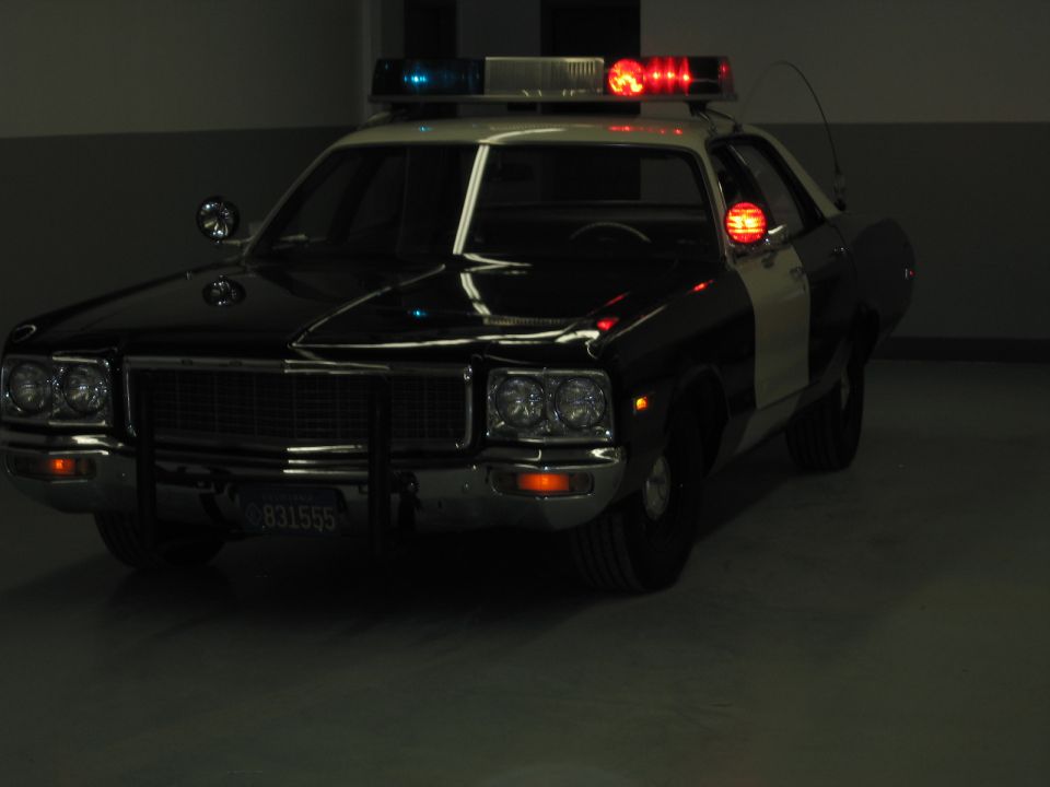 Dimly lit black-and-white Dodge police car with quad headlights and lights flashing