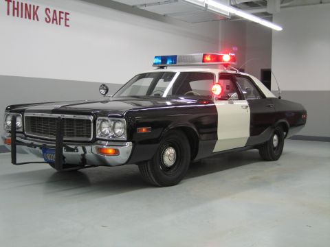 1991 Ford LTD Crown Victoria - black-and-white Dodge police car with chrome push bumper and lit beacons sits below a wall reading THINK SAFE.