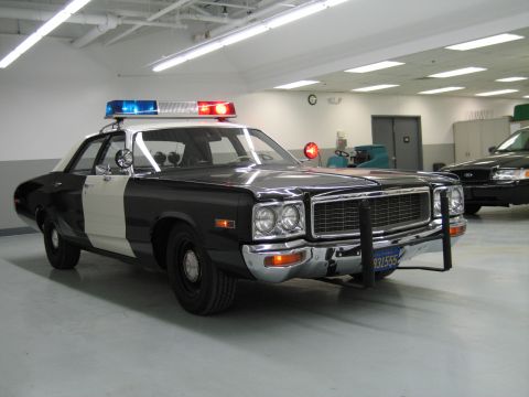 1991 Ford LTD Crown Victoria - black-and-white Dodge police car with chrome grille and push bumper sits in an indoor shop with another car behind.