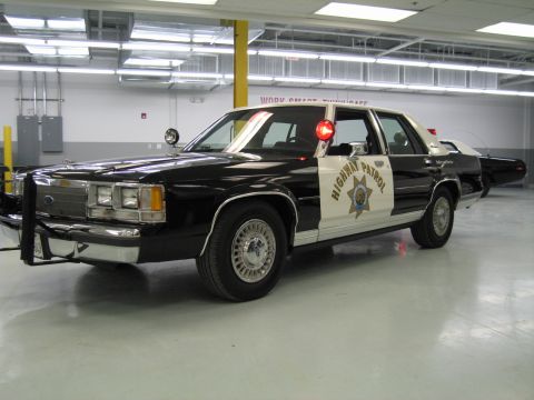 1991 Ford LTD Crown Victoria - black-and-white Ford CHP sedan with HIGHWAY PATROL star door emblem and push bumper parked in a garage.