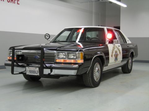 1991 Ford LTD Crown Victoria - Front three-quarter view of a black-and-white CHP Ford sedan with push bar, spotlight, and gold star door emblem.