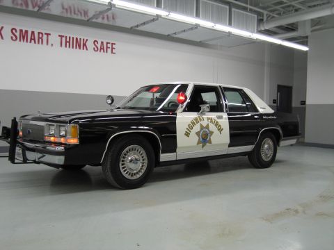 1991 Ford LTD Crown Victoria - Side profile of a black-and-white California Highway Patrol Ford sedan in a shop under a Work Smart Think Safe sign.