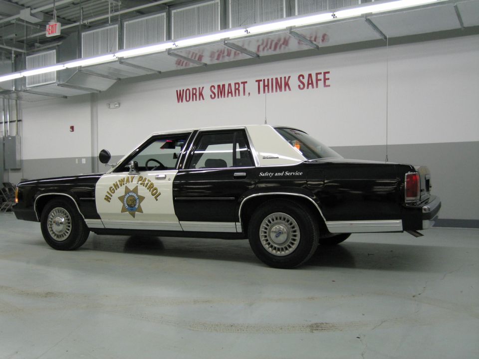 Rear-quarter side view of black-and-white CHP Ford sedan in shop