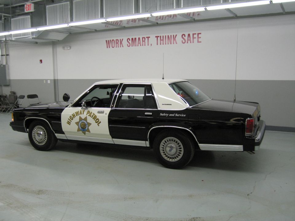 Driver side rear-quarter of black-and-white CHP Ford sedan in shop