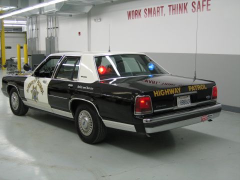 1991 Ford LTD Crown Victoria - Rear three-quarter view of a black-and-white CHP Ford sedan with illuminated red and blue emergency lights.
