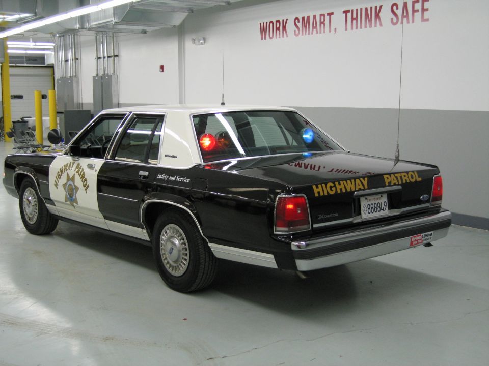 Rear-quarter black-and-white CHP Ford sedan with lit emergency lights