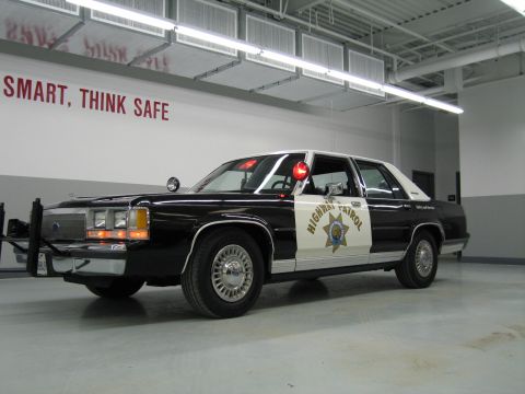 1991 Ford LTD Crown Victoria - Front three-quarter view of a black-and-white CHP Ford sedan with push bar and gold star door decal in a shop.