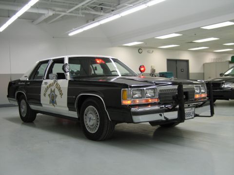 1991 Ford LTD Crown Victoria - Front three-quarter view of a black-and-white CHP Ford sedan with push bar; another dark sedan sits behind it.