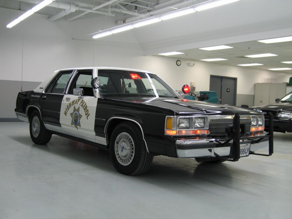 Front-quarter black-and-white CHP Ford sedan with push bar and spotlight indoors