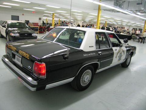 1991 Ford LTD Crown Victoria - Rear three-quarter view of a black-and-white CHP Ford sedan in a busy shop with people and other vehicles behind.