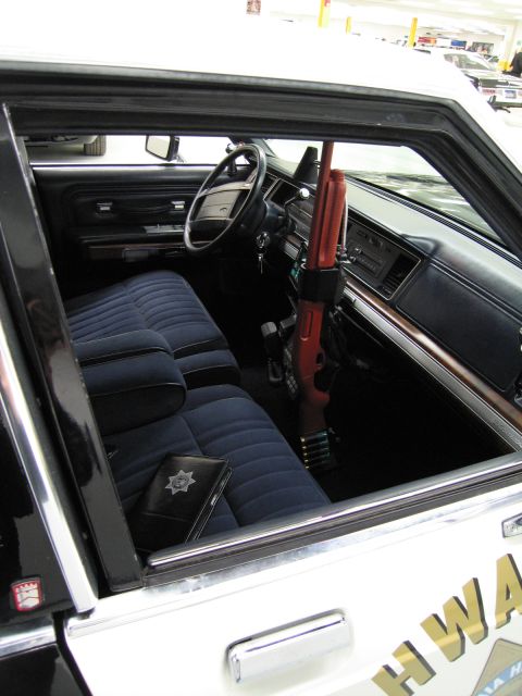 1991 Ford LTD Crown Victoria - Interior of a police car through the open door showing a blue bench seat, dashboard, and a shotgun mounted upright.