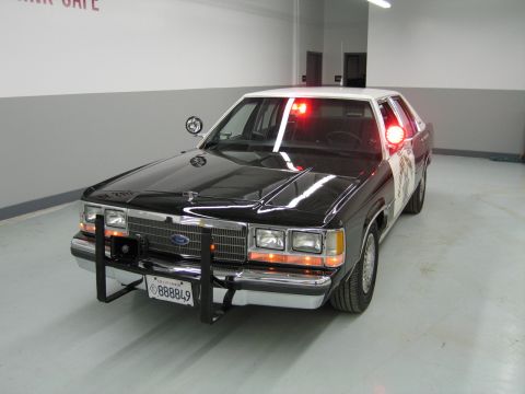 1991 Ford LTD Crown Victoria - black-and-white Ford police interceptor sedan with a front push bar and red roof lights sits in a shop bay with a California plate.