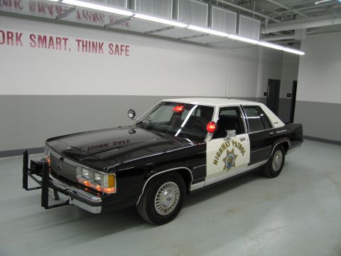 1991 Ford LTD Crown Victoria - black-and-white CHP Ford sedan with star door emblem and roof lights parked in a garage beneath a 'Work Smart, Think Safe' wall sign.