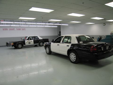 1991 Ford LTD Crown Victoria - Two black-and-white police cars in a garage: an older CHP sedan at left and a newer Ford Crown Victoria-style sedan in the foreground.