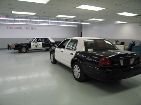 1991 Ford LTD Crown Victoria - Rear three-quarter view of a black-and-white police sedan parked next to an older CHP cruiser under a 'Work Smart, Think Safe' sign.