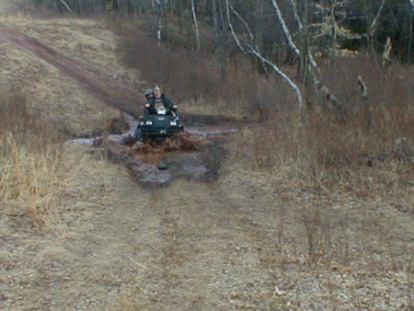 Rider on a dark ATV plowing through a muddy water hole on a rural trail surrounded by dry grass and bare trees