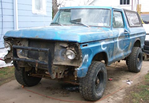 1978 Ford Bronco - A blue 1978 Ford Bronco sits in a yard, showing signs of wear and tear, with its front end partially dismantled.