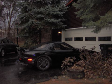 1993 Mazda RX-7 - A black Mazda RX-7 is parked in front of a garage on a cloudy day.