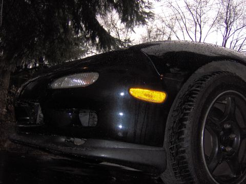 1993 Mazda RX-7 - The front corner of a black 1993 Mazda RX-7 is visible, with a wet finish and yellow turn signal illuminated.