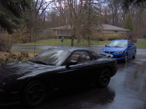1993 Mazda RX-7 - Two cars, a black 1993 Mazda RX-7 and a blue sedan, parked on a wet driveway.