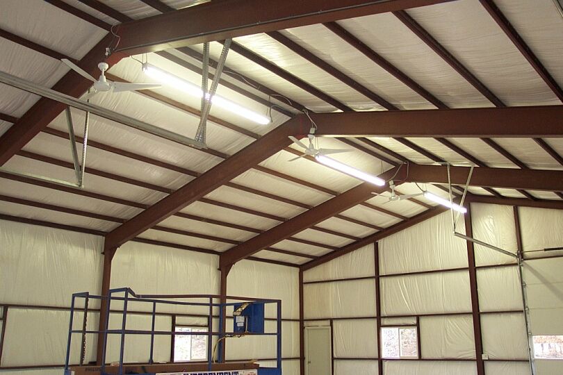 Steel shop building interior with red I-beams, ceiling fans, fluorescent lights and blue lift