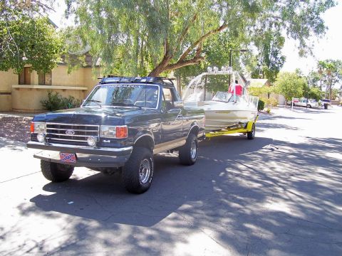 1991 Ford Bronco - A dark blue Ford F-Series truck tows a boat on a trailer down a residential street.